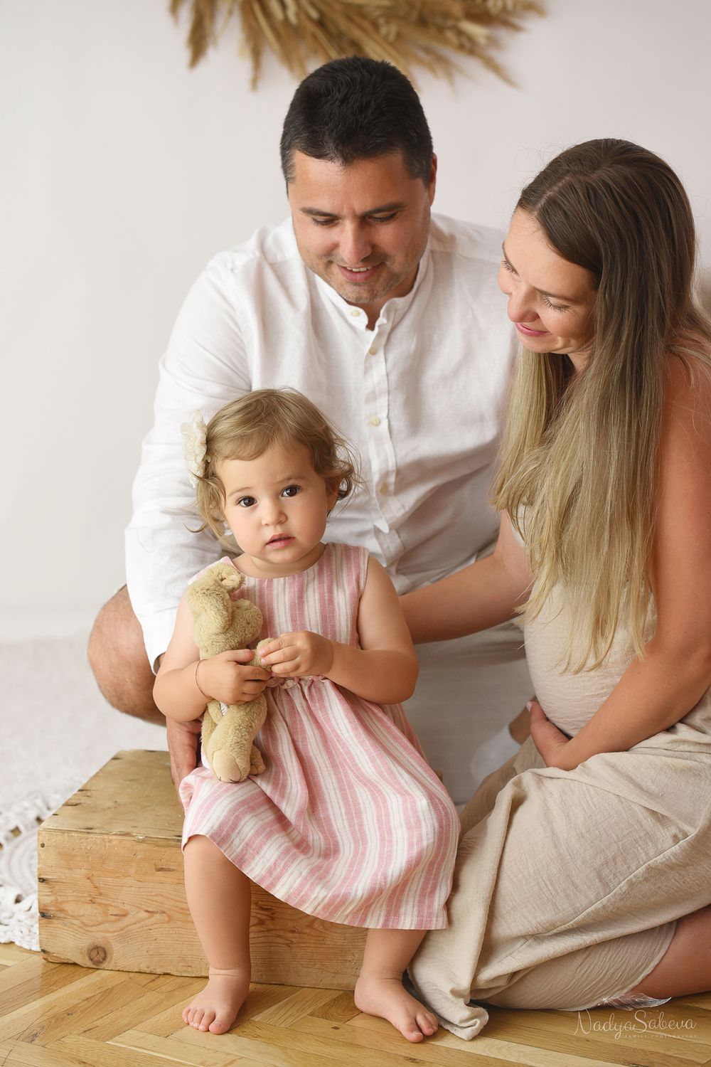 Family sits together on wooden floor as toddler in pink dress holds a stuffed toy.