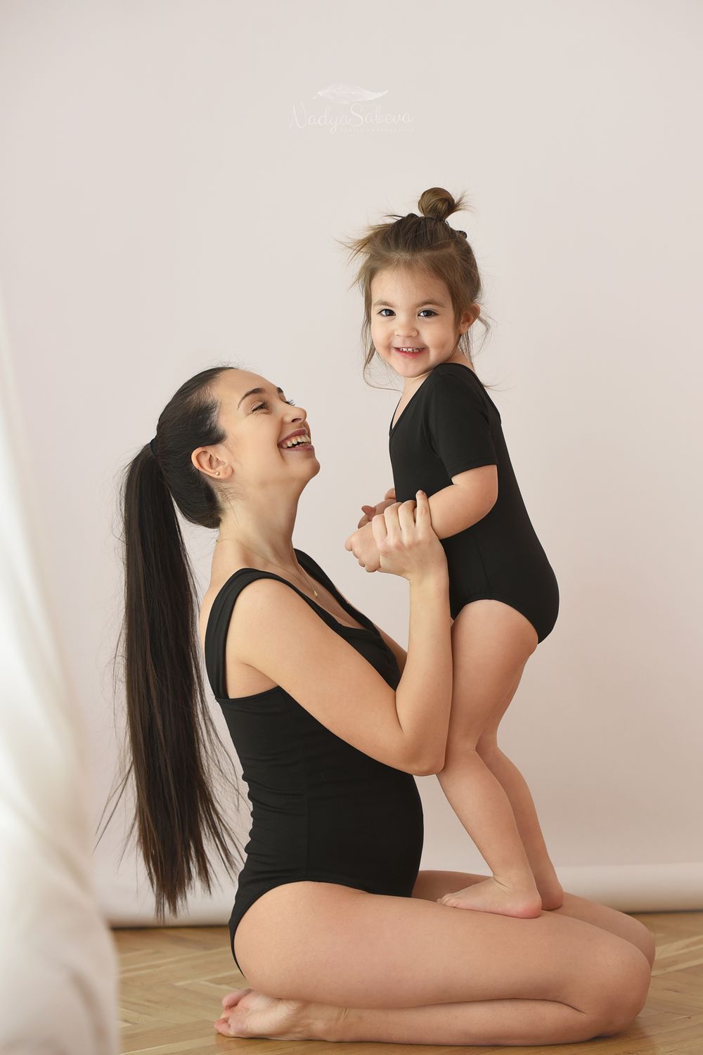 Two figures in black leotards pose together on a wooden floor against a light background for a matching portrait.