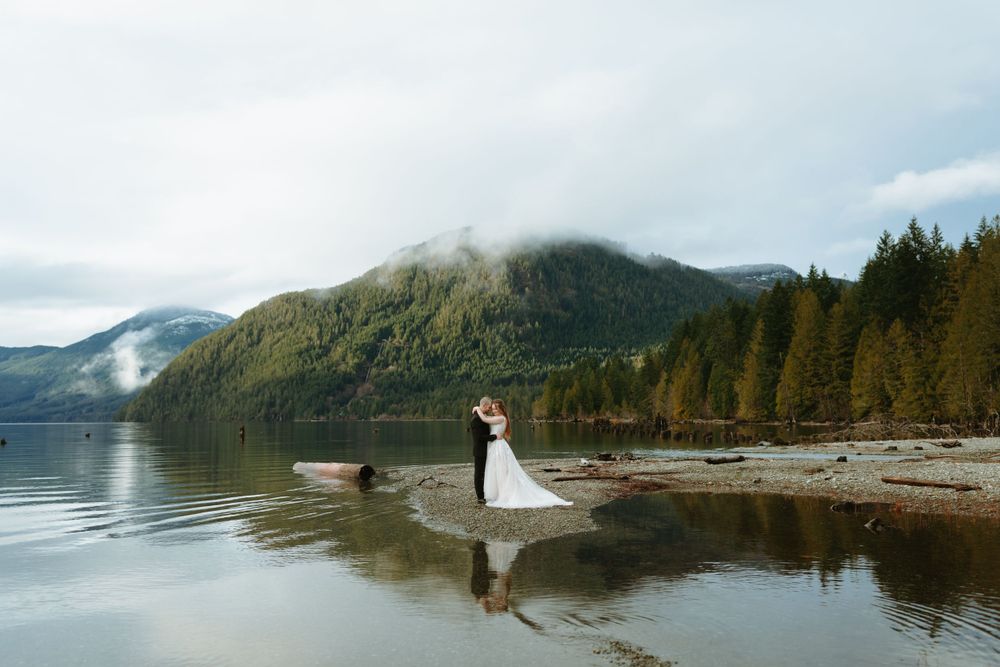 A scenic wedding photo on a misty mountain lake shore with evergreen forests and mountains reflecting in calm waters.