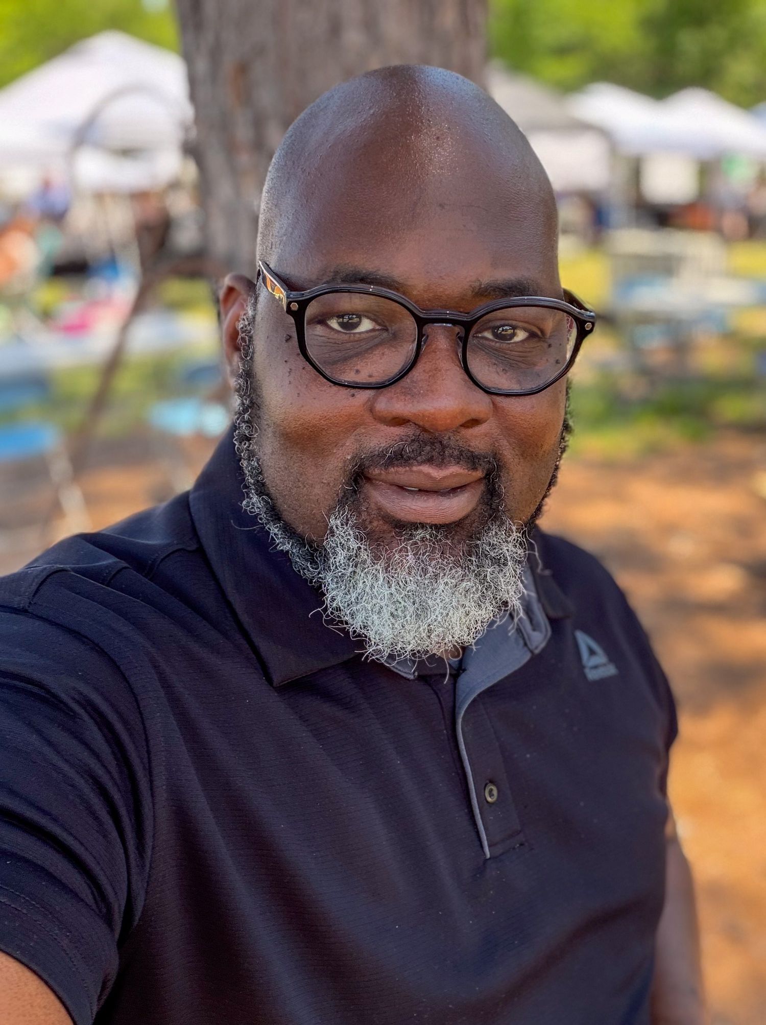 Bald man with gray beard wearing glasses and black polo shirt outdoors.