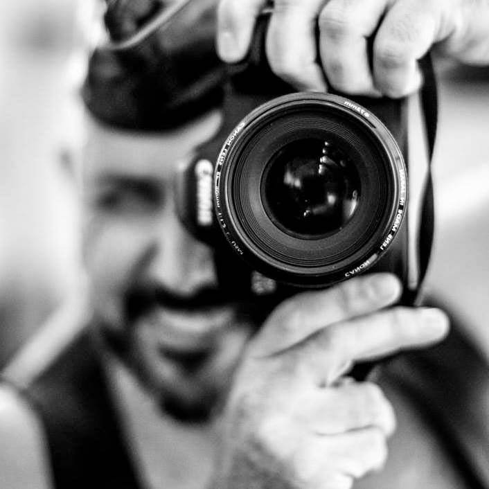 Smiling man holds Canon DSLR camera close to lens in black and white photography portrait.