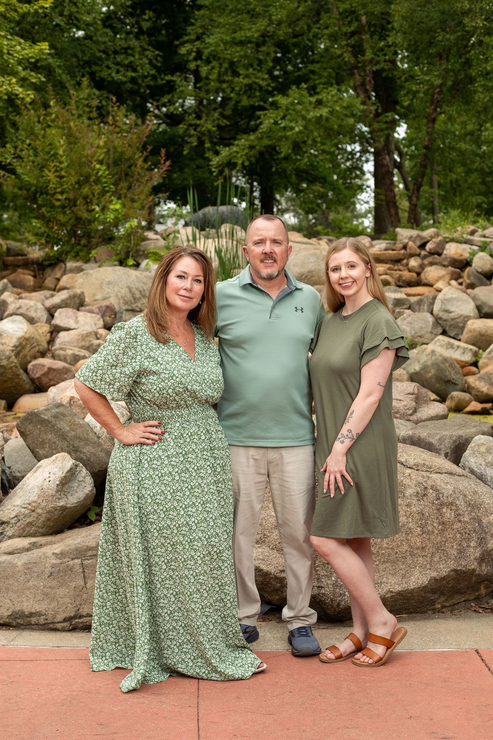 Three adults in green outfits pose together outdoors near a rocky landscape with lush trees in the background.