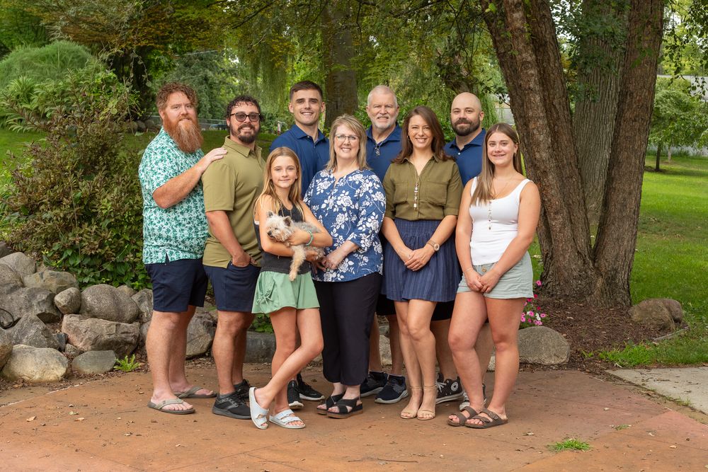 Large family group portrait outdoors, smiling adults posing together near stone wall and green trees.