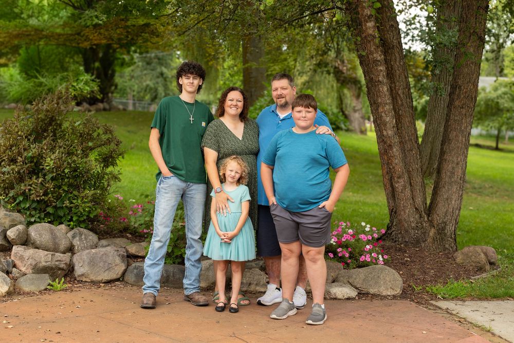 Family of five posing outdoors on a brick path surrounded by lush green trees and garden landscaping.