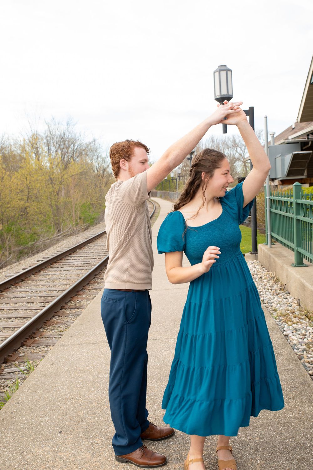 A couple in casual wear dance together on a train platform with railroad tracks visible in the background.