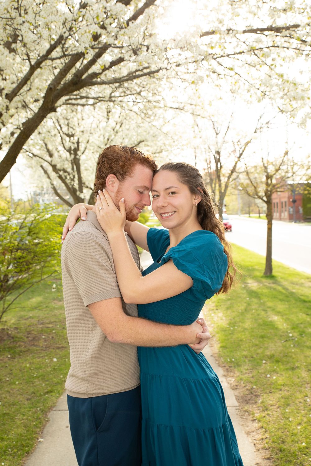 A couple embraces amid blooming cherry blossom trees on a spring day in a grassy park setting.