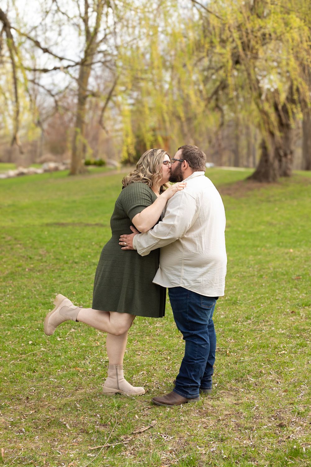 A couple embraces in spring with blooming yellow trees in the background and green grass below.