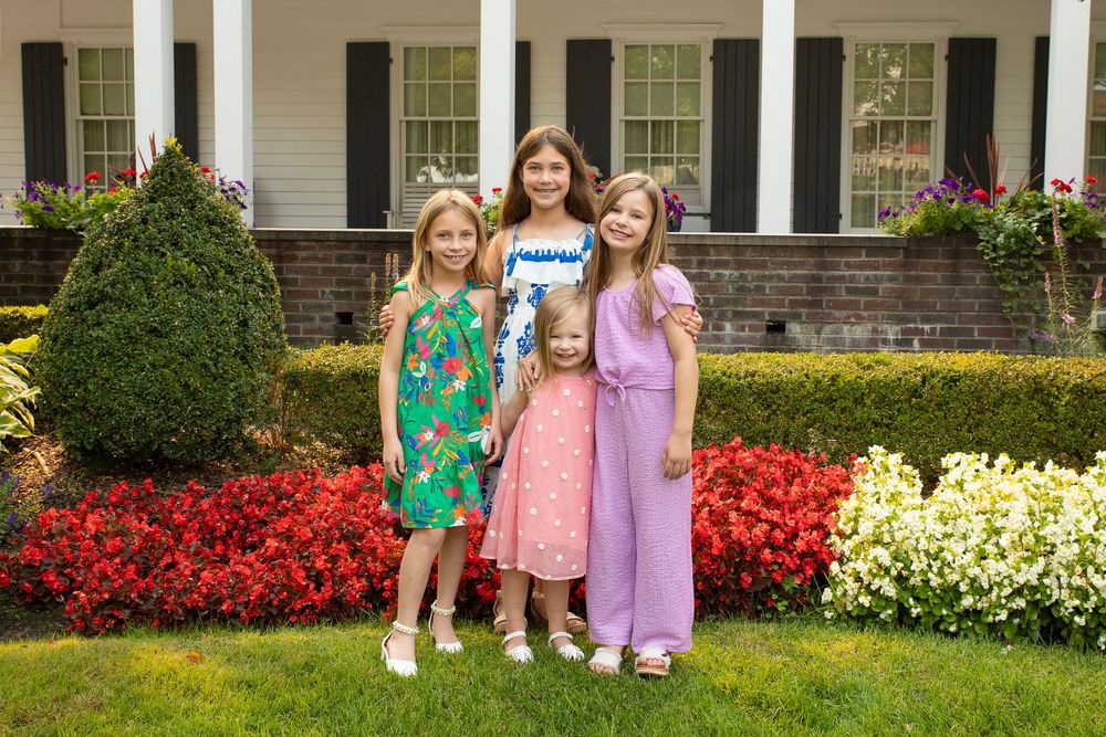 Three children stand in front of a white colonial style house with colorful flower gardens and landscaping.