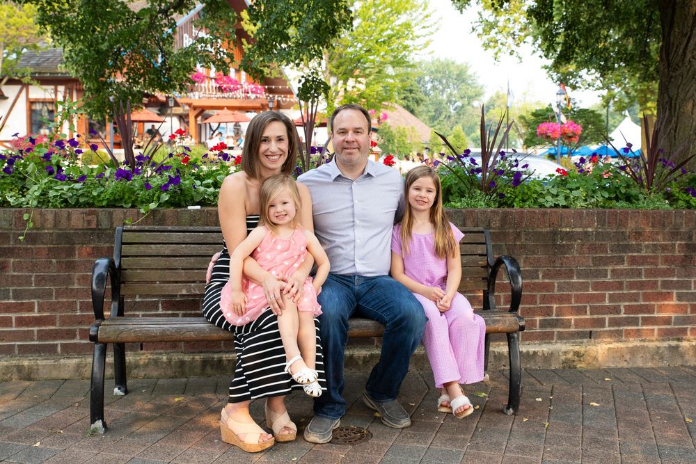 A family sits together outdoors on a brick wall surrounded by lush greenery and blooming flowers.