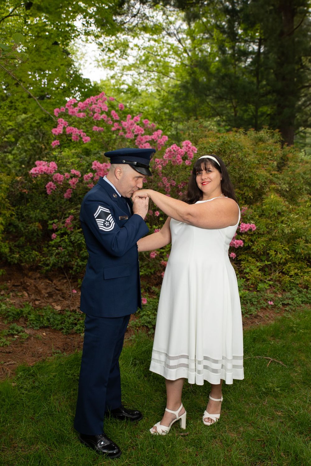 A couple shares a romantic moment in a garden with blooming pink azalea bushes, wearing dress whites and military uniform.