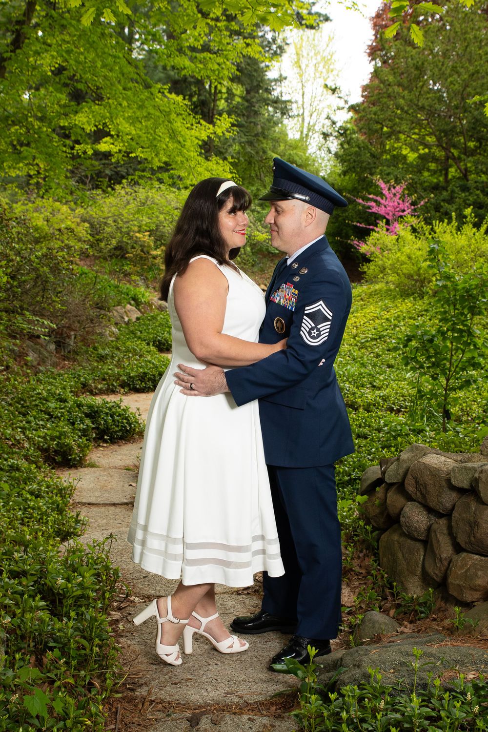 A couple in white dress and air force uniform embrace on a garden path surrounded by lush greenery and blooming flowers.