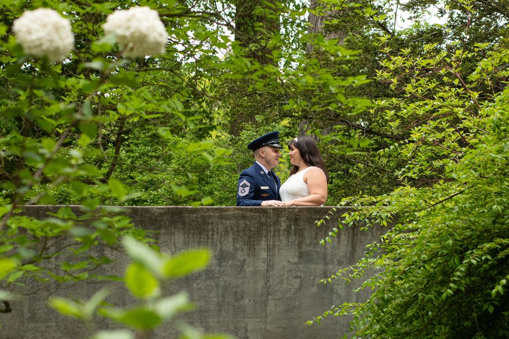 Two people sharing an intimate moment on a concrete wall surrounded by lush green foliage in a garden setting.