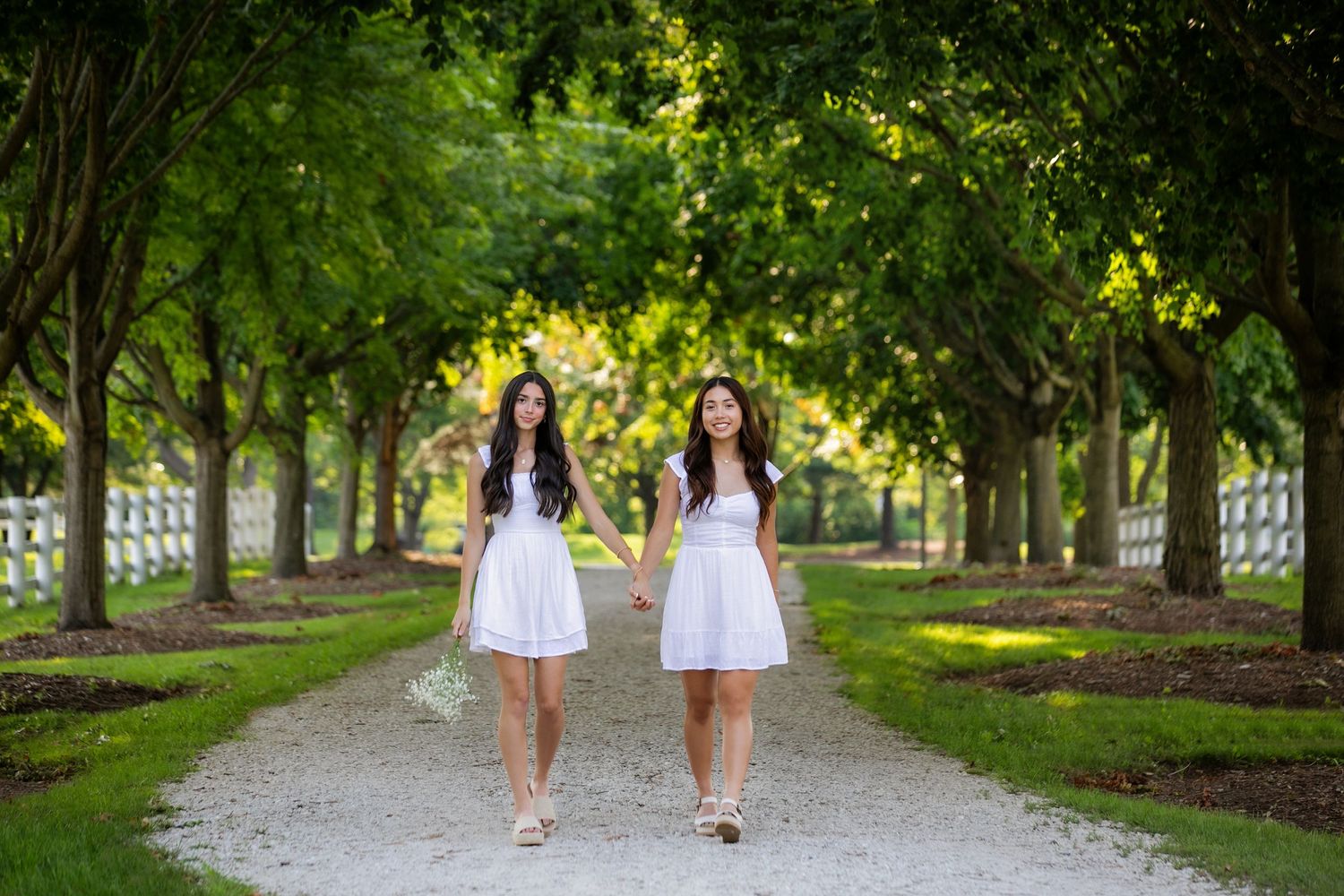 Two best friends senior session in matching white dresses walking down a tree-lined path.