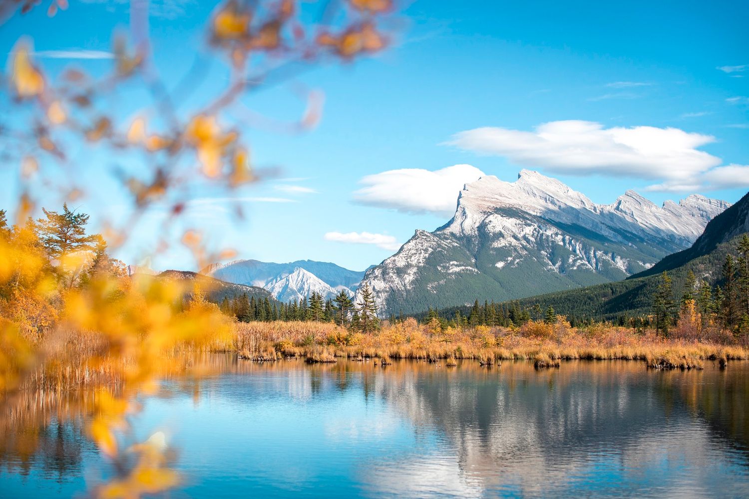 A crystal-clear mountain lake reflects autumn foliage and a majestic peak under a bright blue sky.