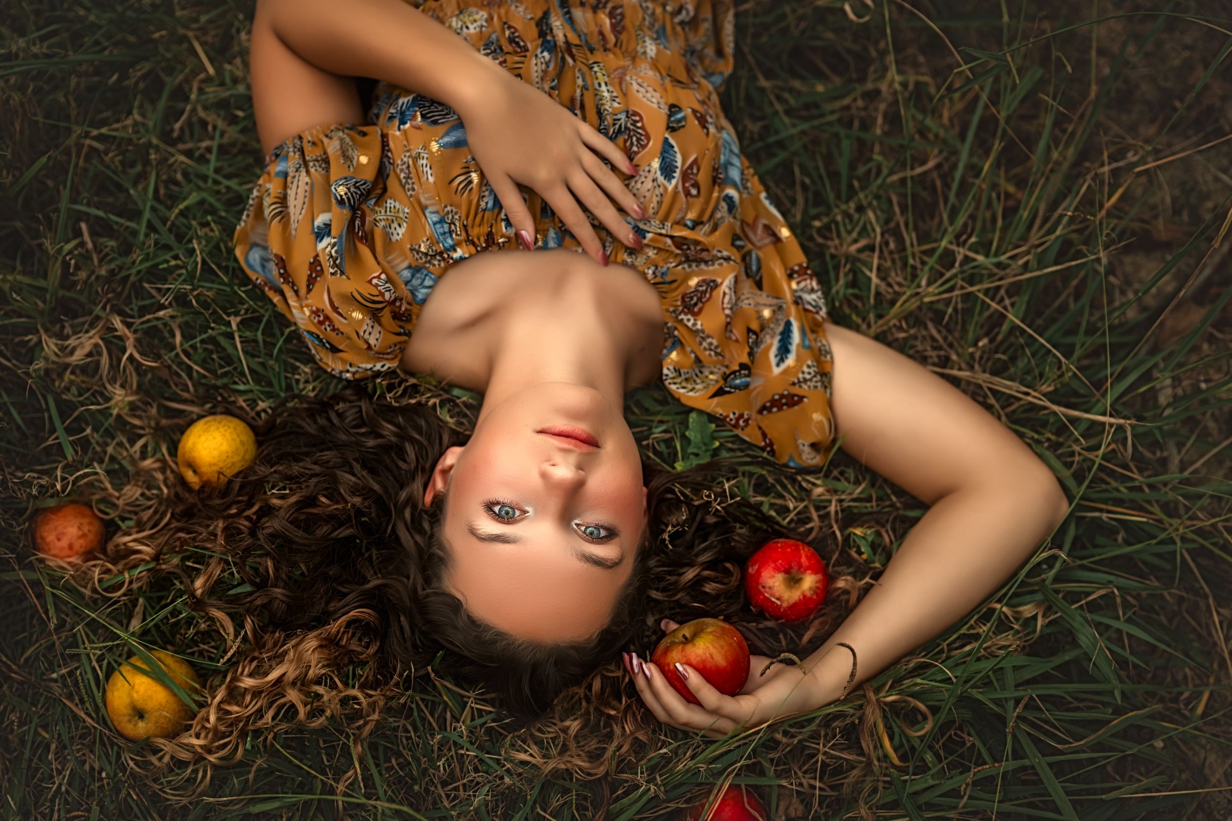 Senior girl lying in grass with scattered red apples on dark ground in artistic portrait series for fine art senior photos.