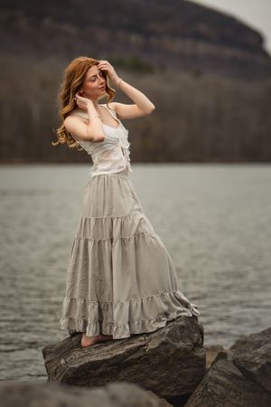 Artistic portrait series showing senior girl in a flowing boho top and skirt posing gracefully on rocks by the water on cloudy day.
