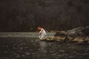 A senior girl in a white dress sits on rocks by a misty lake, creating a moody and ethereal atmosphere.