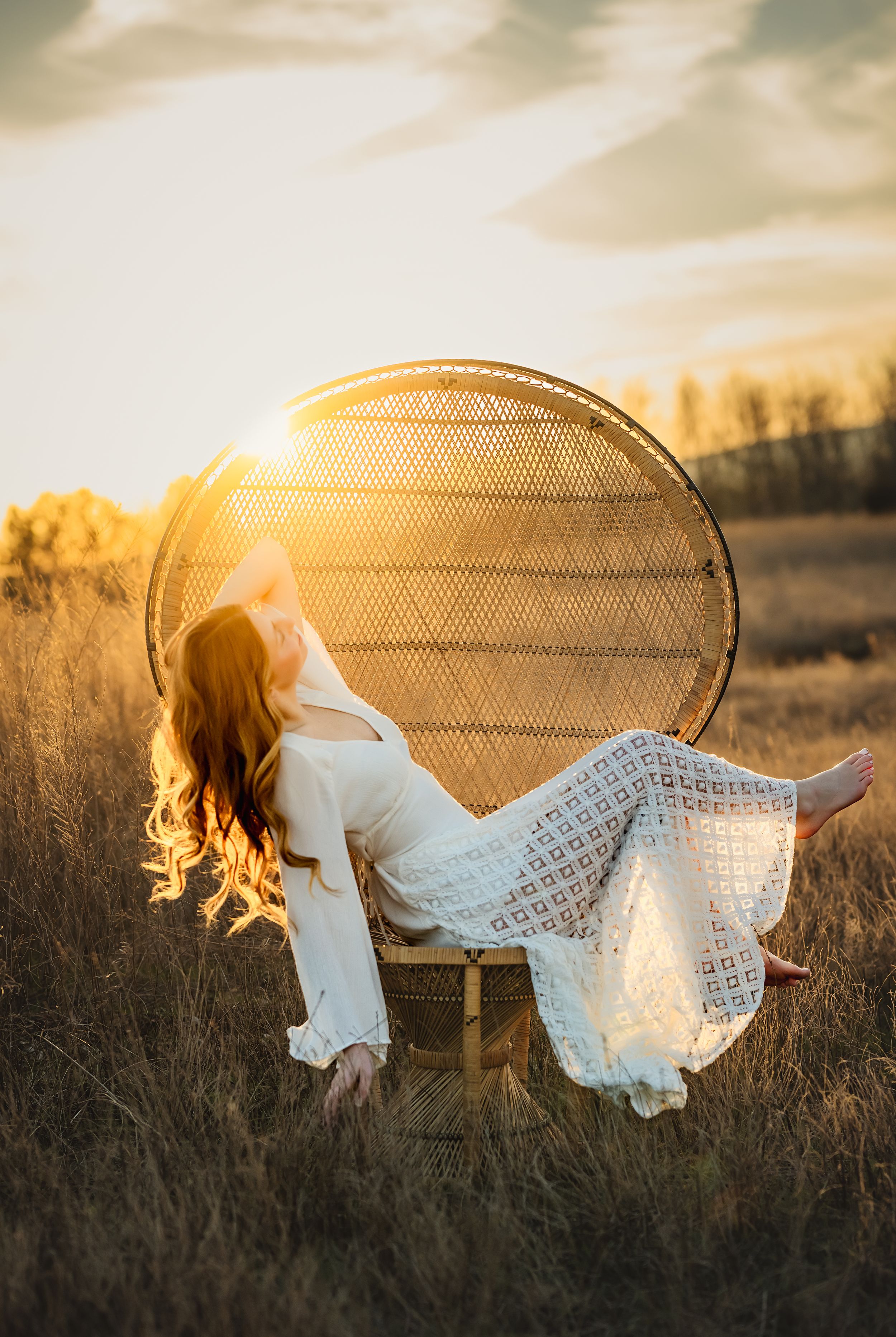 A dreamy golden sunset scene shows a senior girl in a white dress lounging ethereally in a peacock chair in the tall grass.