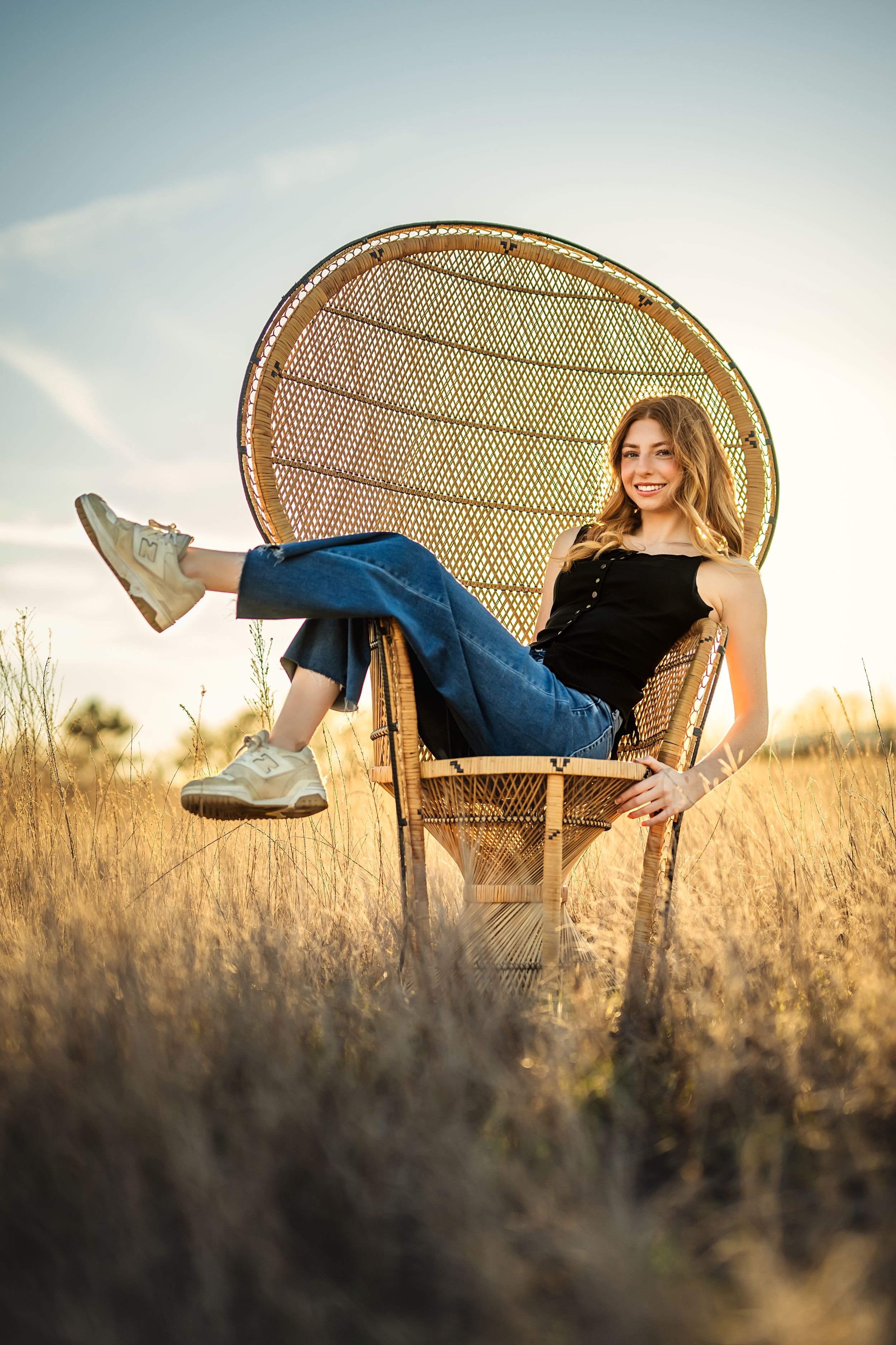 High school senior girl relaxing in a peacock chair while wearing black top and jeans in golden field at sunset for Chattanooga senior photo