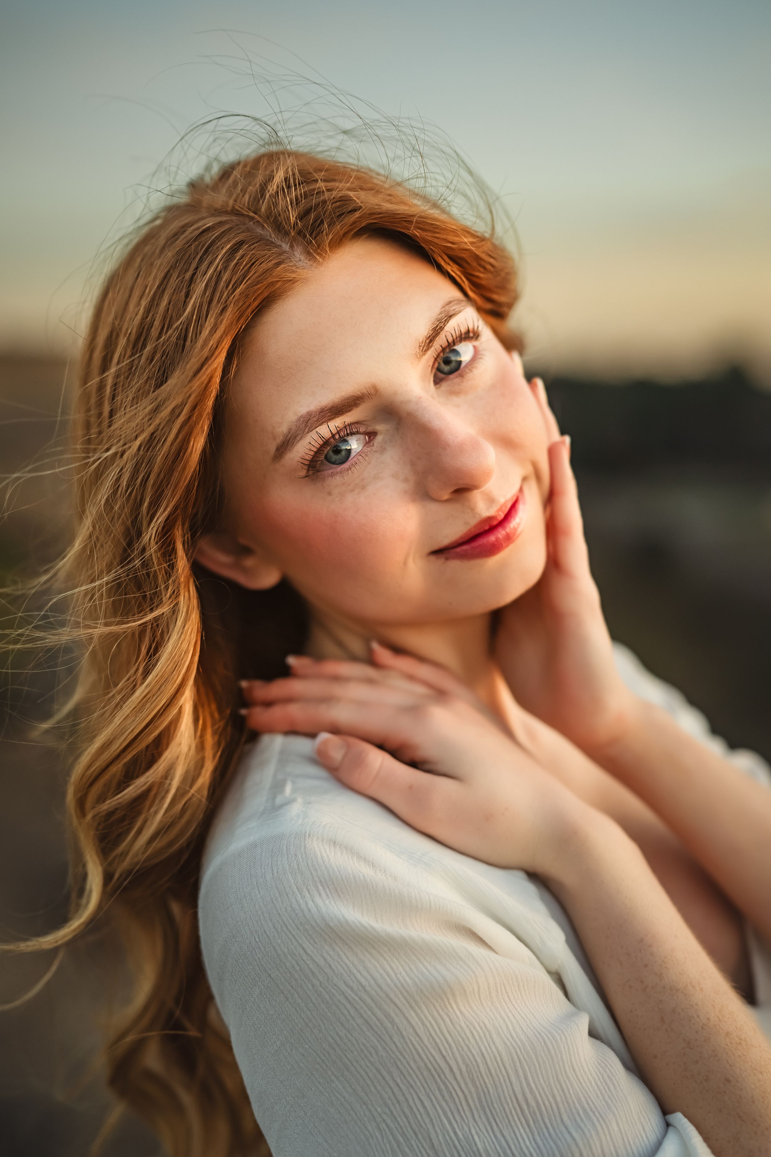 Senior girl with windswept red hair poses for close-up in a white dress at sunset for senior photo session.