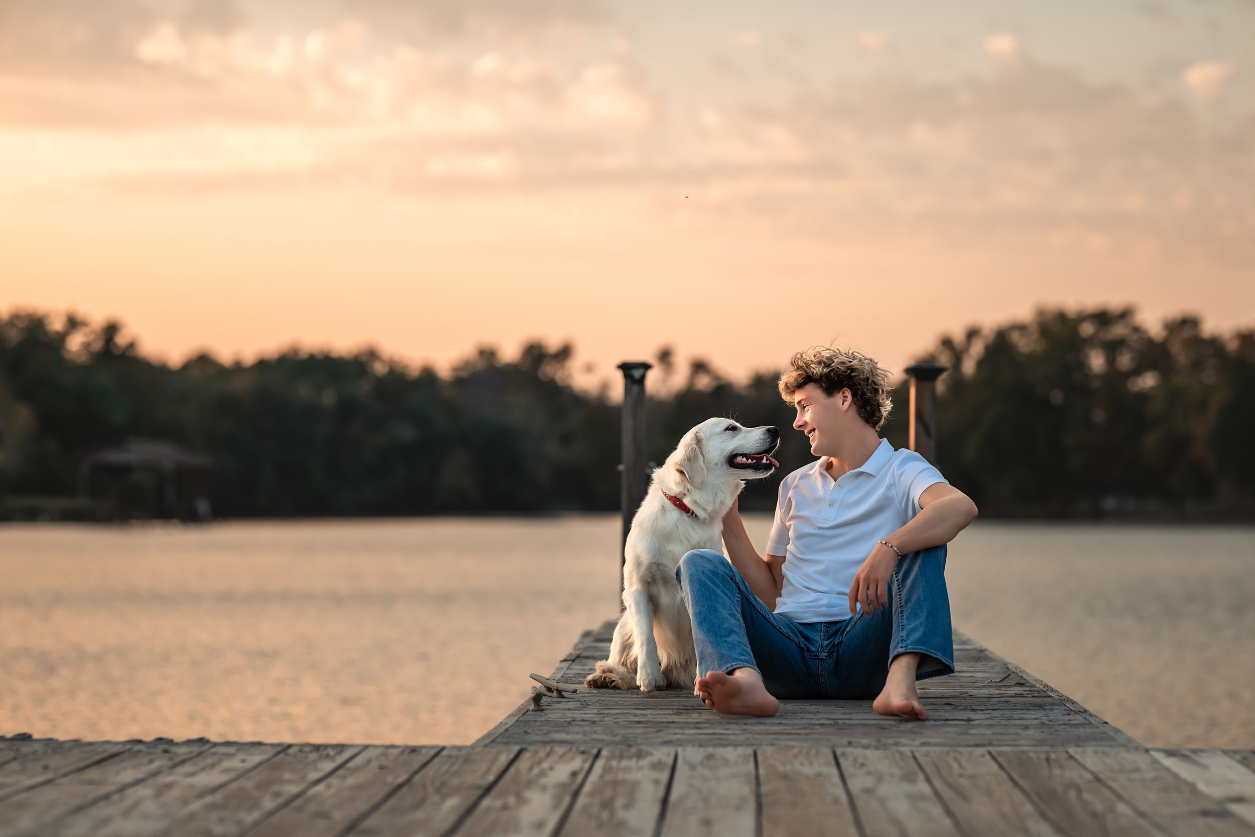 Senior boy relaxing on wooden dock by water with his dog during peaceful sunset with orange sky and reflection.