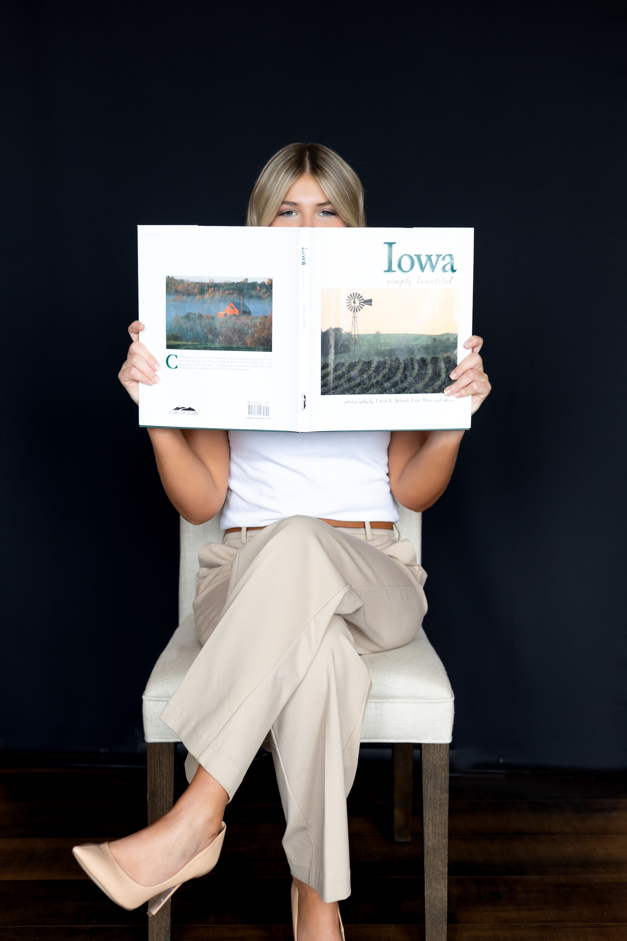 Someone wearing khaki pants and a white top sits holding an Iowa travel magazine on a chair against a dark wall.