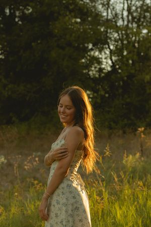 Woman in floral sundress smiling with eyes closed in a sunlit meadow at golden hour.