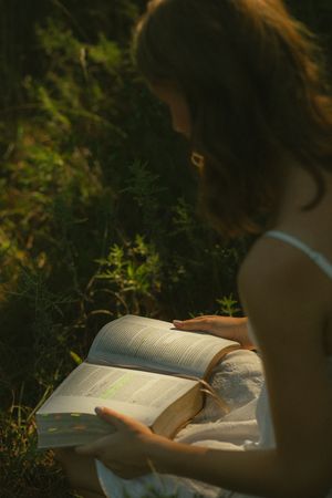 Young woman in white dress reading a large open book outdoors in dappled sunlight among green foliage.
