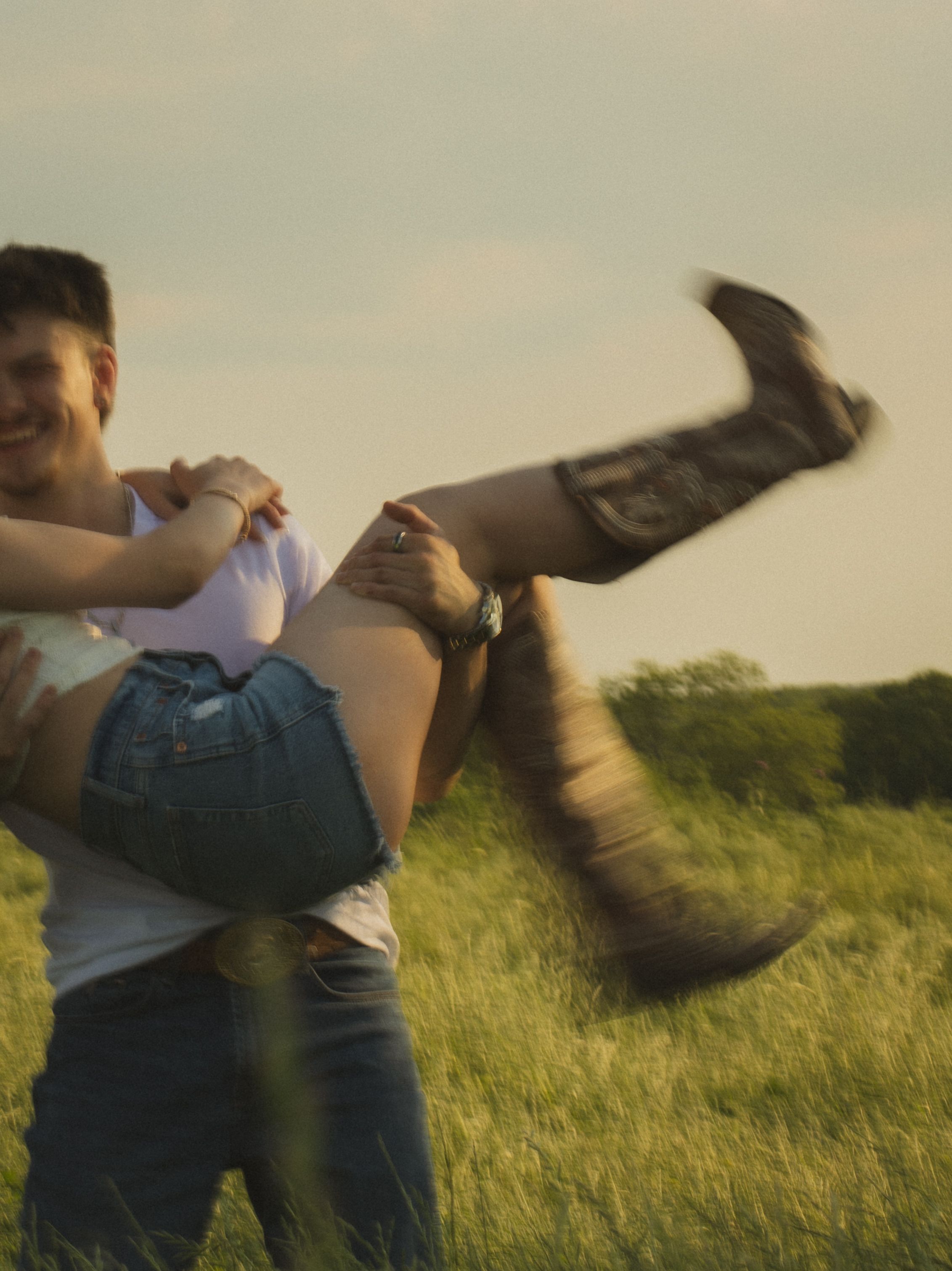 Couple laughing in sunny green field, man lifting woman wearing cowboy boots and denim shorts outdoors.
