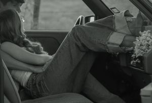 Black and white photo of a couple sitting together inside a vintage car with wildflowers.