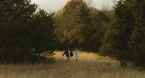 Two people running hand-in-hand through golden tall grass field surrounded by evergreen trees in autumn light.