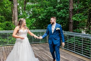 Couple in wedding attire walks hand in hand across wooden bridge in forest setting.
