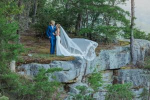 A couple poses dramatically on a rocky cliff edge with flowing veil in the wind.