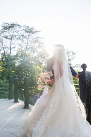A bride in a flowing white dress and veil holds a pink bouquet against sunlit trees on an outdoor deck.
