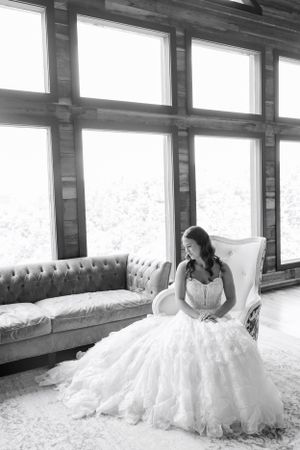 A bride in a ballgown sits on a white chair near large windows in black and white.