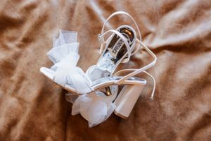 A pair of elegant white bridal heels with straps and bows laid on brown fabric.