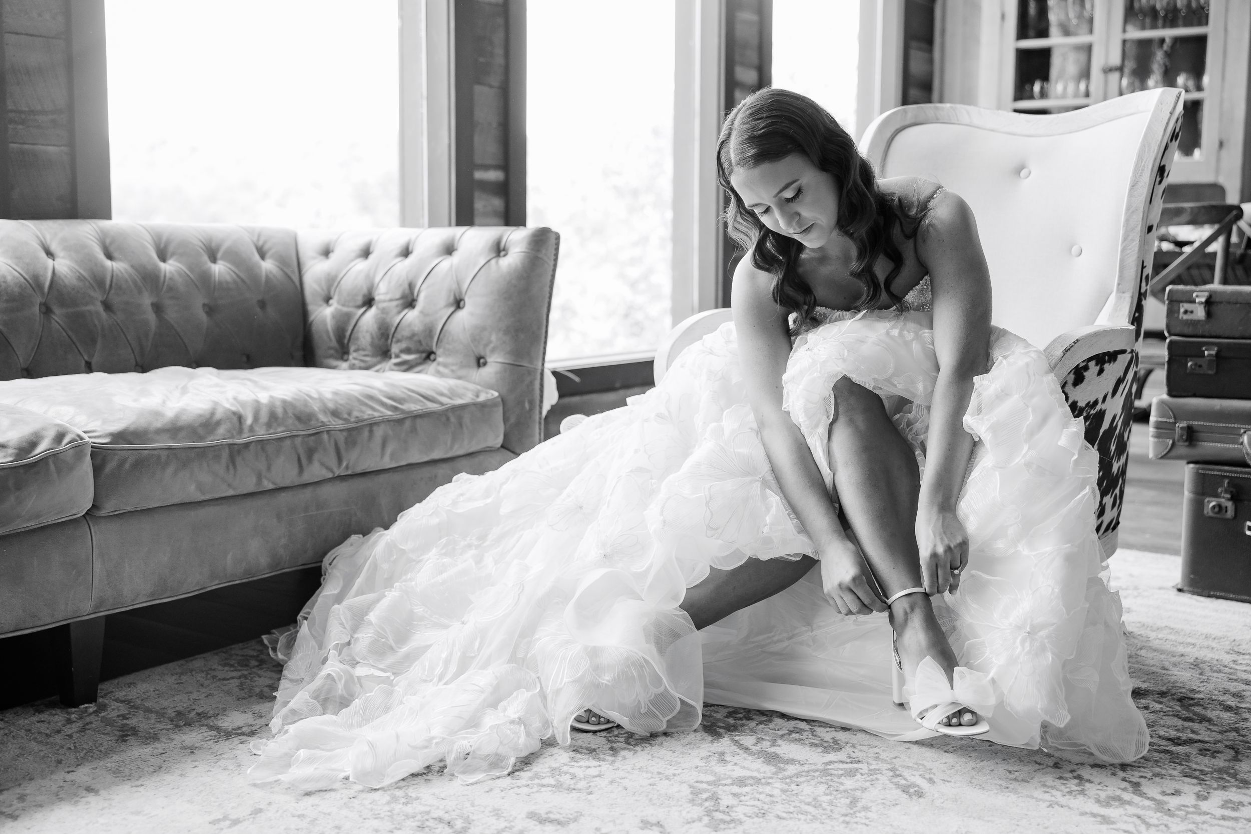 A bride in a wedding dress puts on her shoes while seated in an elegant room.