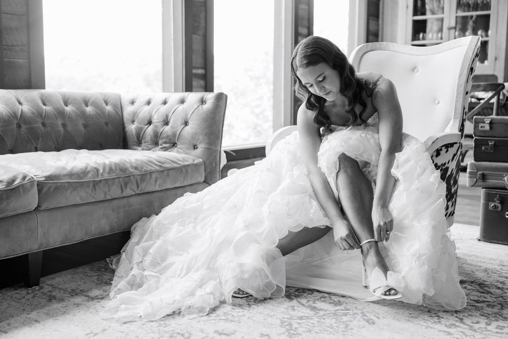A bride in a wedding dress puts on her shoes while seated in an elegant room.