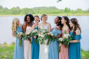 Bridesmaids in various blue dresses hold lush green bouquets by waterfront.