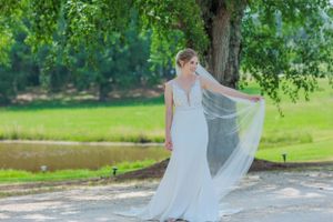 Bride in flowing white dress poses with veil by lake under tree.