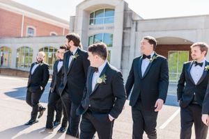 Groomsmen in tuxedos walking outside church building on sunny day.