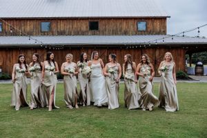 Bridal party in champagne colored dresses poses together outside rustic venue.