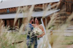 A romantic wedding portrait through tall grass with string lights and barn in background.
