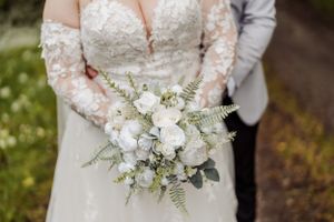 Elegant white wedding bouquet with roses and greenery held against lace dress.