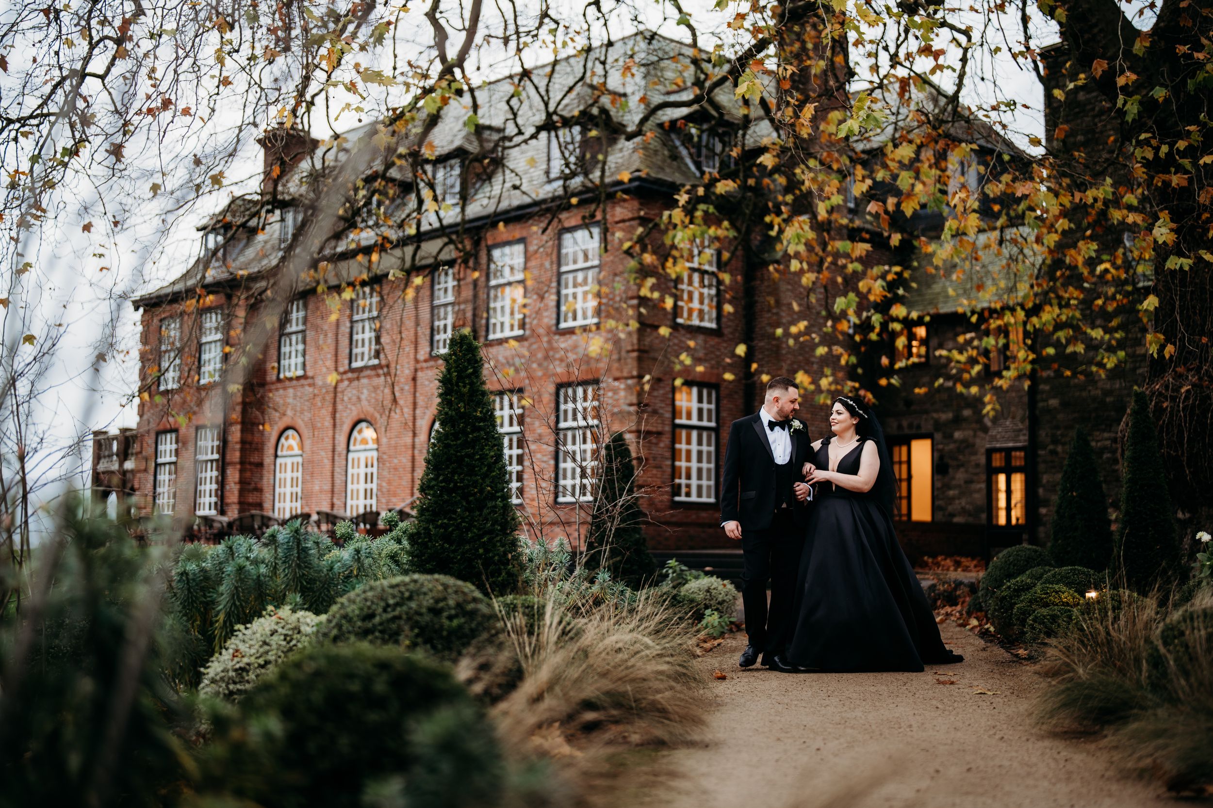 A couple stands together on autumn grounds of a grand Victorian brick manor house with weeping willow trees.