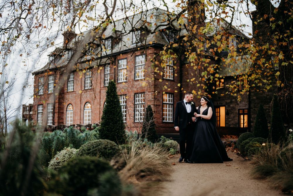 A couple stands together on autumn grounds of a grand Victorian brick manor house with weeping willow trees.