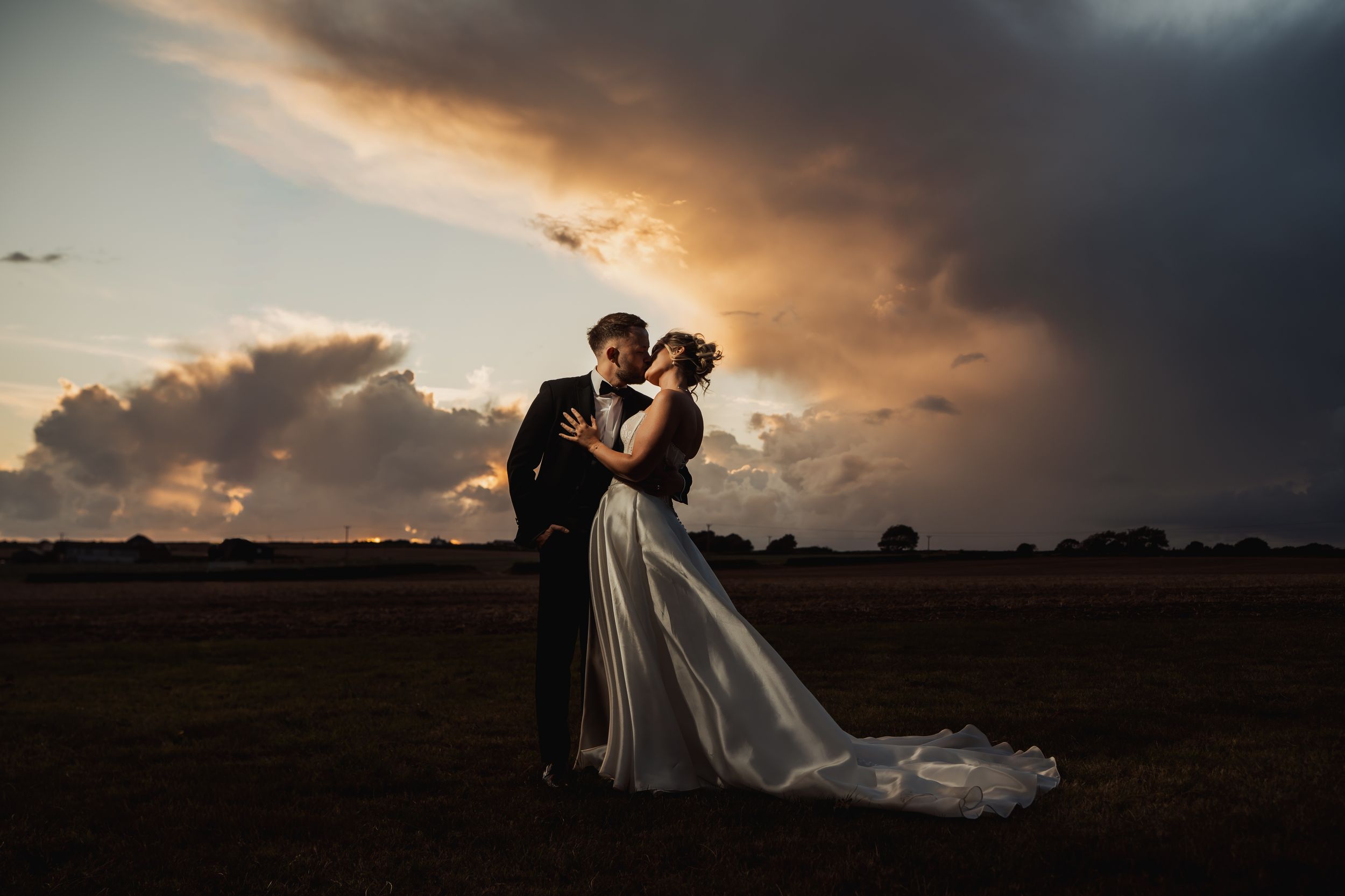 A couple embraces in a romantic silhouette against a dramatic sunset sky in an open field.