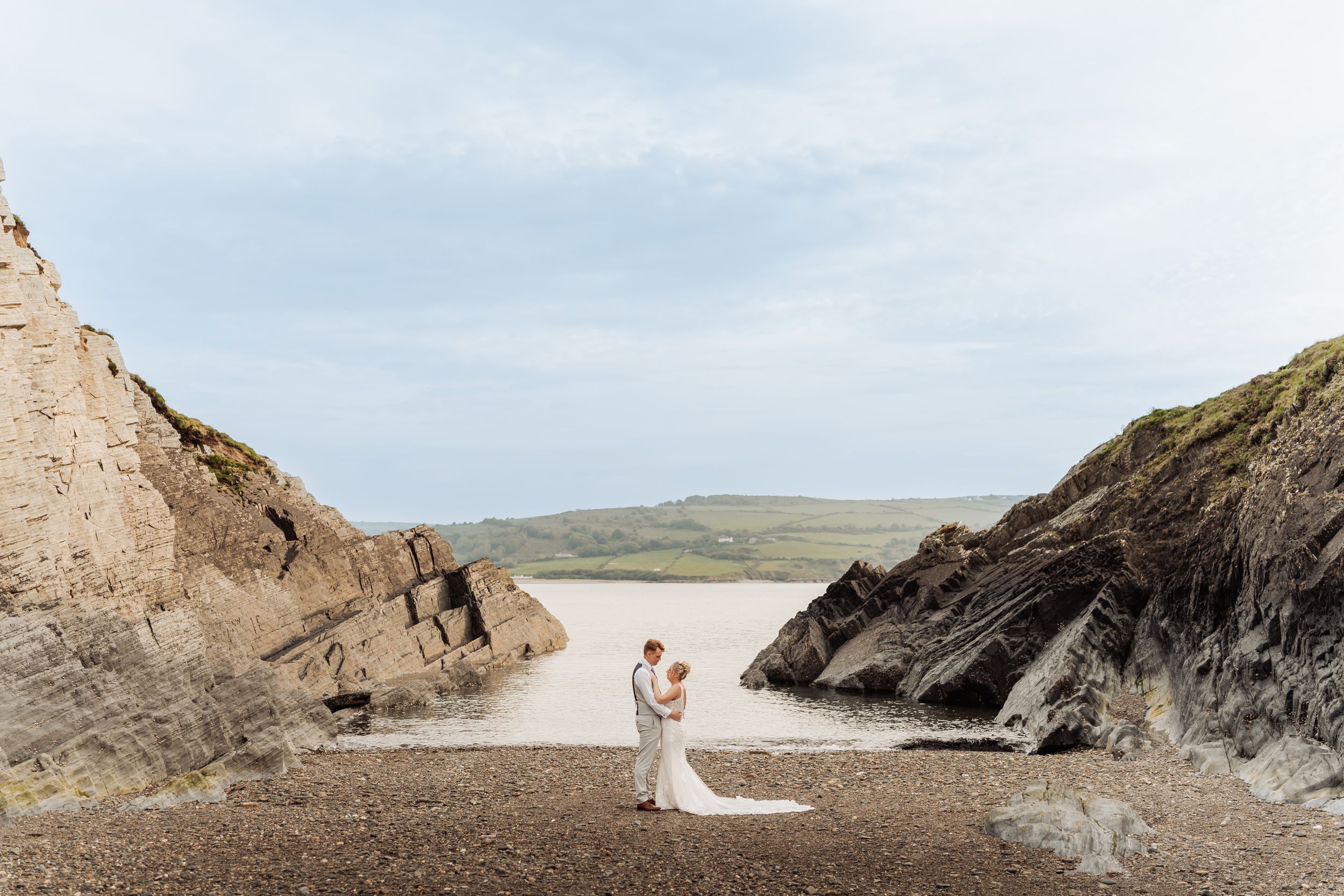 A couple stands between two dramatic rocky cliffs overlooking a scenic beach and ocean view on a cloudy day.