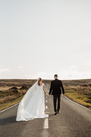 Newlyweds walking down empty road with flowing white dress and dramatic sky.