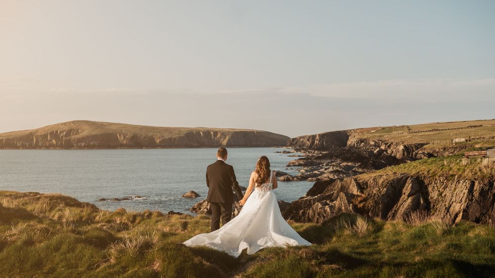 Two figures stand on dramatic coastal cliffs overlooking a serene ocean bay during golden hour in a scenic landscape.